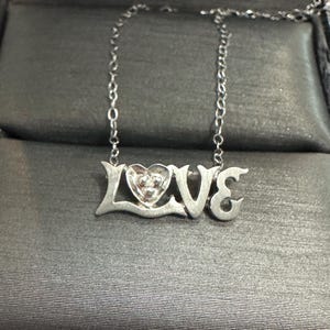 Könnte beinhalten: Eine silberne Halskette mit dem Wort "LOVE" als Anhänger. Das "O" ist herzförmig mit einem kleinen Edelstein in der Mitte. Die Halskette hat eine silberne Kette.