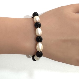 May include: A bracelet featuring alternating black and white beads. The black beads are round, while the white beads are oval-shaped. The bracelet is worn on a wrist, showcasing the jewelry's design and color contrast.