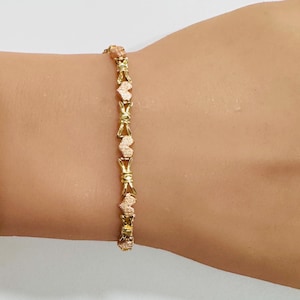 May include: A gold bracelet featuring a pattern of heart-shaped and X-shaped links. The hearts have a textured, rose-gold finish, contrasting with the smooth gold X-links. The bracelet is displayed on a wrist, highlighting its intricate design.