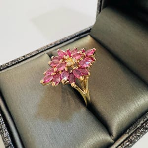 May include: A gold ring featuring a cluster of pink marquise-cut gemstones arranged in a floral design. The ring has a delicate, detailed setting and is presented in a dark gray jewelry box.
