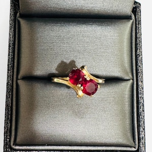 May include: A gold bypass ring featuring two round, faceted, deep red gemstones set side-by-side. The band has a smooth, polished finish and curves around the stones, which are held in place by small metal prongs in a classic, elegant arrangement.