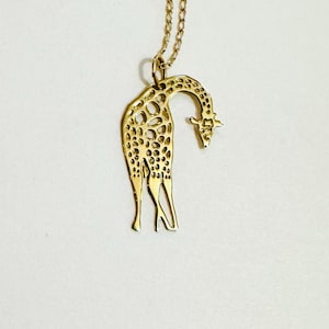 Puede incluir: Un collar con colgante dorado con la silueta de una jirafa. La jirafa tiene un diseño texturizado y abierto con cuello y patas largos. La cadena del collar también es dorada y delicada.