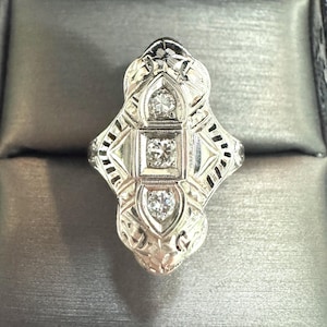 Op de afbeelding: Een gedetailleerde ring in Art Deco-stijl met drie prominente diamanten gezet in een filigraan ontwerp. De ring heeft een langwerpige, symmetrische vorm met ingewikkeld metaalwerk en een gepolijste afwerking. De diamanten zijn gezet in vierkante en ovale zettingen.