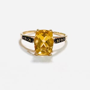 Könnte beinhalten: Ein goldener Ring mit einem großen, kissenförmigen gelben Citrin, der von vier Krappen gehalten wird. Die Ringschiene ist aus Gold gefertigt und weist auf beiden Seiten des Mittelsteins zwei dunkle, texturierte Metallakzente auf.