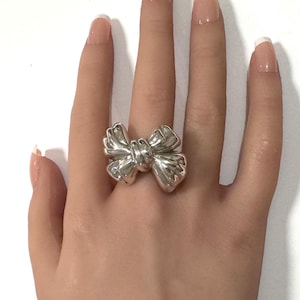 May include: A polished silver ring featuring a prominent three-dimensional bow design. The metallic surface is smooth and reflective, showcasing a sculptural knot at the center with two symmetrical loops extending outward on each side of the band.