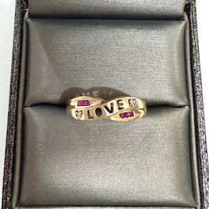 May include: A gold-toned ring featuring the word LOVE in raised lettering. The band has an infinity-style shape and is accented with two rectangular red gemstones and two small, clear, round-cut stones set on either side of the central text.