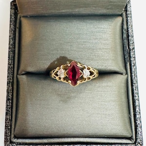 May include: A gold ring featuring a marquise-cut red gemstone, with two smaller white gemstones either side. The ring has an ornate, filigree design. It is presented in a grey velvet-lined jewellery box.
