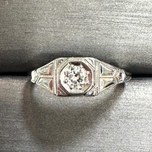 May include: A silver ring with an octagonal center setting, featuring a sparkling round diamond. The ring has intricate filigree detailing on the shoulders and around the central stone, creating a vintage aesthetic.