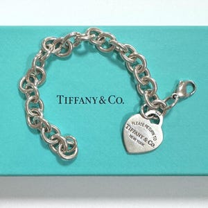 May include: A silver Tiffany & Co. bracelet with a heart-shaped tag that reads "PLEASE RETURN TO TIFFANY & CO. NEW YORK." The bracelet is on a teal blue surface. The bracelet has a lobster clasp.