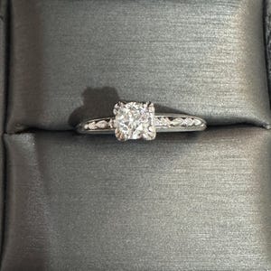 May include: A silver engagement ring with a large, square-cut diamond center stone. The band features smaller accent stones. The ring is set against a gray, textured background, likely inside a ring box.