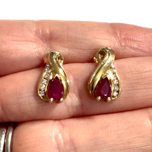 May include: A pair of gold stud earrings featuring a central pear-shaped red ruby gemstone surrounded by a curved gold setting accented with small, clear, round-cut white diamonds along the side of each earring, creating a teardrop shape.
