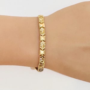 May include: A gold-colored bracelet with an intricate design. The bracelet is made of multiple rectangular and circular segments, creating a textured appearance. The bracelet is worn on a wrist.
