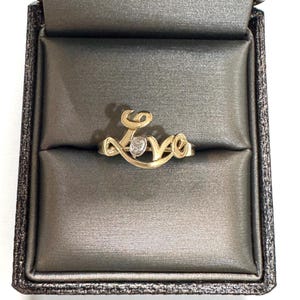 May include: A gold ring featuring the word LOVE in cursive script, with a small, round, clear stone set in the center of the letter O. The ring is displayed inside a dark gray, padded jewelry box with a satin-like finish and a textured exterior.