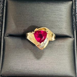 May include: A gold ring featuring a large, triangular, vibrant red gemstone. The ring has a distinctive design with a swirl detail and small, rectangular, clear stones. The ring is presented in a black jewellery box.