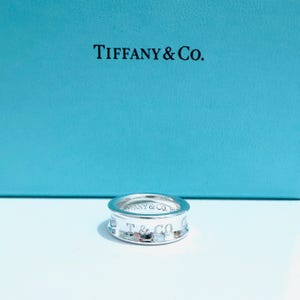 May include: A silver ring with the engraved text "Tiffany & Co." is displayed against a light blue background. The ring is circular with a flat band and a polished finish. The brand name is visible on the ring.
