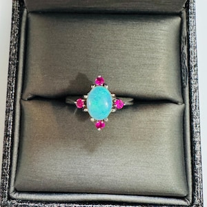 May include: A silver ring featuring a large, oval, light blue opal gemstone, surrounded by smaller, vibrant pink gemstones. The ring is set against a dark gray, textured background, showcasing the jewelry's design and color contrast.