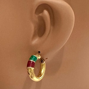 May include: A gold hoop earring featuring two distinct enamel bands in vibrant green and deep red. The earring has a polished metallic finish and a circular design that curves to form a classic hoop shape, secured through the earlobe.