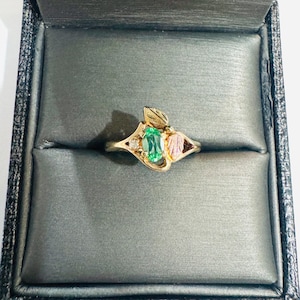 May include: A gold ring with a green oval-cut gemstone, accented with small diamonds and leaf-shaped details. The ring is set in a black box with a gray interior, showcasing the jewelry's design and craftsmanship.