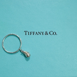 Könnte beinhalten: Ein silberner Ring mit einem Tropfenanhänger, vor einem blauen Hintergrund von Tiffany & Co. Das Band des Rings ist ein dünner Kreis, und der Anhänger hat eine glatte, abgerundete Form. Das Tiffany & Co. Logo ist schwarz gedruckt.