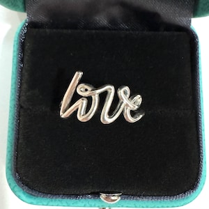 May include: A silver-colored ring in the shape of the word "love" is displayed inside a black velvet jewelry box with a teal exterior. The ring is in a cursive font.