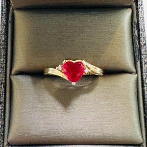 May include: A gold ring with a heart-shaped, deep red gemstone as the centerpiece. The ring features small accent stones and is set in a decorative band. The ring is displayed in a jewelry box.