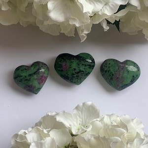 Könnte beinhalten: Drei herzförmige Steine in Grün und Pink mit schwarzen Flecken. Die Steine sind in einer Reihe auf einer weißen Oberfläche angeordnet.