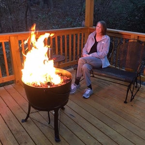 May include: A woman sits on a wooden deck with a fire pit in the centre. The fire pit is made of metal and has a round base with three legs. The fire pit is filled with burning wood and embers. The woman is wearing a grey jacket and jeans. The deck is made of wooden planks and is surrounded by a wooden railing.