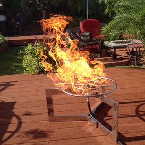 May include: A stainless steel fire pit with a circular burner and a square base. The fire pit is on a wooden deck and is surrounded by flames.