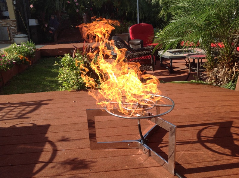 May include: A stainless steel fire pit with a circular burner and a square base. The fire pit is on a wooden deck and is surrounded by flames.