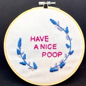 Può includere: Un cerchio da ricamo bianco con un motivo floreale rosa e blu e il testo "HAVE A NICE POOP" in filo rosa.