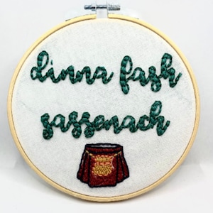 May include: Embroidered hoop art with green text that reads "dinna fash sassenach" and a red embroidered kilt with gold accents.