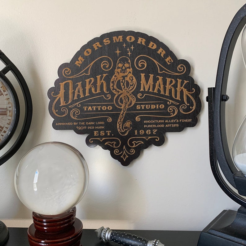 Puede incluir: Un letrero de madera negro con letras doradas que dice "Morsmordre Dark Mark Tattoo Studio" con un gr&aacute;fico de serpiente y cr&aacute;neo. El letrero tambi&eacute;n incluye el texto "Aprobado por el Se&ntilde;or Oscuro 1200 por marca Est. 1967 Knockturn Alley's Finest Pureblood Artists."