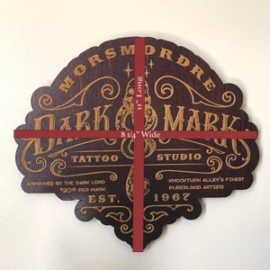 Puede incluir: Letrero de madera marr&oacute;n oscuro con letras doradas que dice "Morsmordre Dark Mark Tattoo Studio" con el texto "Aprobado por el Se&ntilde;or Oscuro 2000 &euro; por marca Est. 1967 Knockturn Alley's Finest Pureblood Artists". El letrero mide 21 cm de ancho y 29 cm de largo.