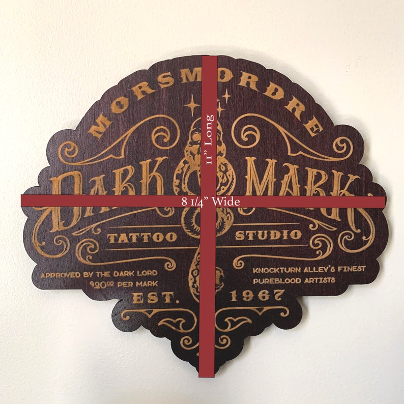 Puede incluir: Letrero de madera marr&oacute;n oscuro con letras doradas que dice "Morsmordre Dark Mark Tattoo Studio" con el texto "Aprobado por el Se&ntilde;or Oscuro 2000 &euro; por marca Est. 1967 Knockturn Alley's Finest Pureblood Artists". El letrero mide 21 cm de ancho y 29 cm de largo.