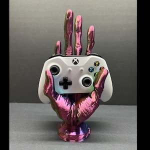 May include: A 3D printed hand holding a white Xbox controller. The hand is painted in a pink and purple iridescent color. The controller has black buttons and a black D-pad.