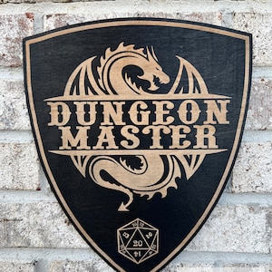 May include: Black wooden shield-shaped sign with the words "DUNGEON MASTER" in a light brown font. A dragon design is above and below the text. A twenty-sided die is at the bottom. The sign is approximately 11.25 inches high and 9.35 inches wide.
