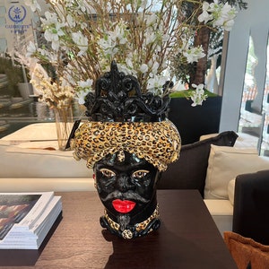 Könnte beinhalten: Schwarze Keramikkopfvase mit Leopardenmuster-Turban und schwarzer Krone. Die Vase hat eine rote Lippe und einen schwarzen Schnurrbart. Weiße Blumen und Zweige sind in der Vase angeordnet. Die Vase steht auf einem dunkelbraunen Tisch.