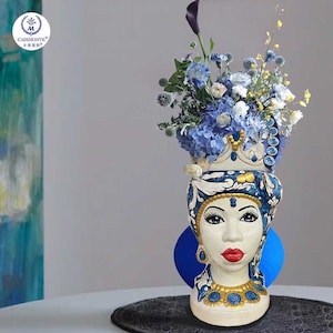 May include: A blue and white ceramic vase shaped like a woman's head with a crown and floral design. The vase is filled with blue and white flowers.