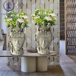 May include: Two off-white ceramic head vases, one male and one female, each adorned with a crown and floral details. The vases hold arrangements of white and green flowers. The vases are on a marble-topped table.