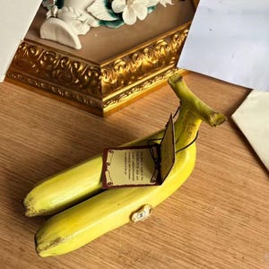 Pode incluir: Uma escultura de banana em cerâmica amarela com um formato curvo realista e um caule no topo. Uma etiqueta de papel retangular está presa com um cordão preto, exibindo o texto impresso: This piece is made in the CAIMONTE style and is hand crafted and painted in keeping with the finest traditions of the art.