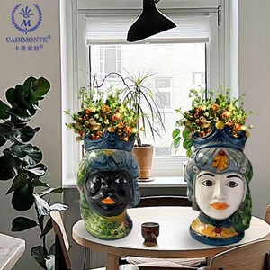 May include: Two ceramic planter pots shaped like human faces. The planter on the left has a black face with a green, blue, and white design. The planter on the right has a white face with a blue, green, and yellow design. Both planters have flowers in them.