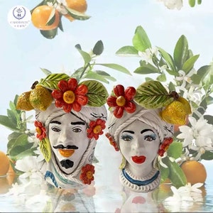 Puede incluir: Dos macetas de cerámica con caras pintadas, flores y limones. La maceta de la izquierda es un hombre con bigote y barba. La maceta de la derecha es una mujer con pestañas largas y labios rojos. Ambas macetas tienen un fondo blanco y están decoradas con detalles rojos, amarillos y verdes.