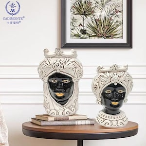 May include: Two ceramic head vases with black faces and gold lips. The vases are adorned with white, ornate headwear and collars. One vase is taller than the other. The vases are on a round, wooden table.