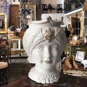 May include: White ceramic vase shaped like a woman's head with a crown and ornate details. The vase is 32 cm tall and 24 cm wide.