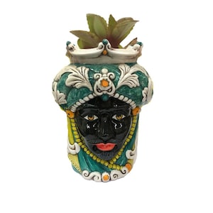 May include: A ceramic planter shaped like a black man's head wearing a green and white turban with a crown. The planter has yellow and orange accents.