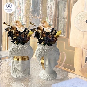 Puede incluir: Dos jarrones de cerámica blanca con forma de cabeza humana con detalles dorados. Los jarrones están llenos de flores secas, incluyendo rosas negras y hojas pintadas de oro.