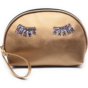 May include: A gold-colored makeup bag with a zipper closure and a black handle. The bag has two sparkly eyelashes on the front.