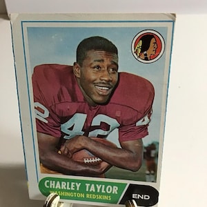 Puede incluir: Una tarjeta de fútbol Topps vintage de la década de 1960 con Charley Taylor, un jugador de los Washington Redskins. La tarjeta está en buen estado con una ligera curva en la esquina inferior derecha. La tarjeta presenta una foto de Taylor con su uniforme, sosteniendo un balón de fútbol. La tarjeta también incluye el logotipo del equipo y el texto "CHARLEY TAYLOR WASHINGTON REDSKINS END".