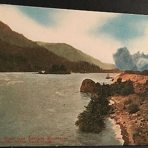 Peut inclure: Une carte postale ancienne représentant un train circulant le long du fleuve Columbia avec les montagnes Cascade en arrière-plan. Le train émet une grande colonne de fumée. Le texte sur la carte postale indique "Columbia River and Cascade Mountains on the North Bank Road."