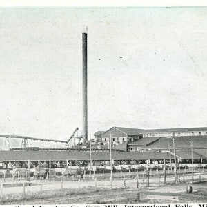 Könnte beinhalten: Ein Schwarzweißfoto eines großen Industriegebäudes mit einem hohen Schornstein. Das Gebäude hat ein langes, niedriges Dach und viele Fenster. Im Vordergrund befinden sich mehrere kleinere Gebäude, und ein Zaun umgibt den gesamten Komplex.